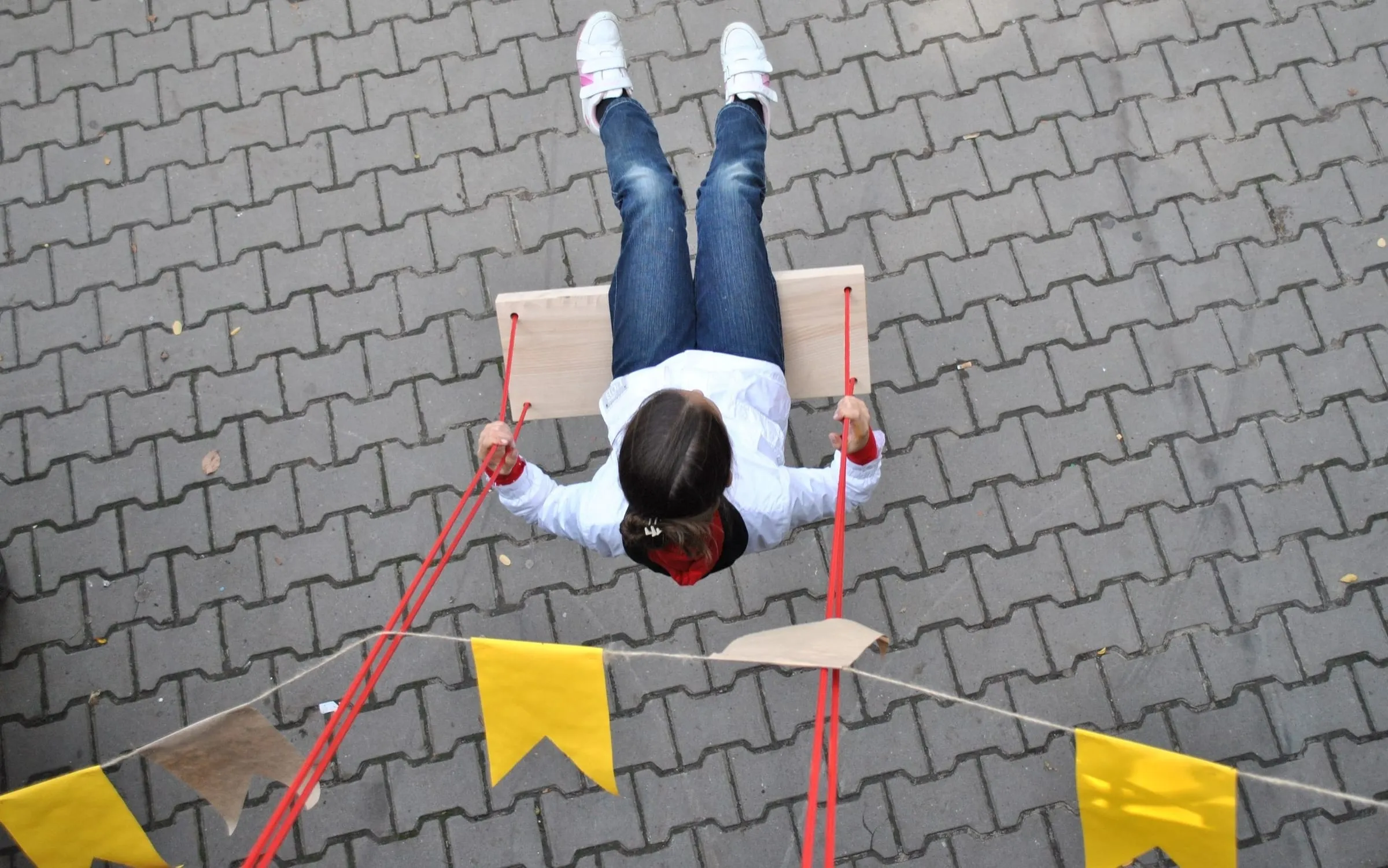 A child on a rope swing above paving stones and yellow festival flags, seen from above